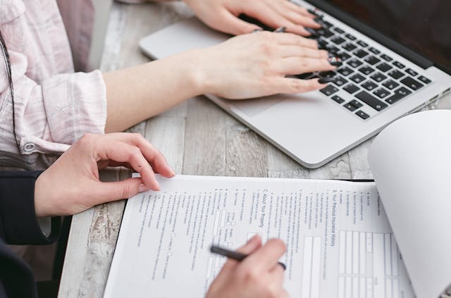 Person holding a pen reviewing a document next to someone using a laptop